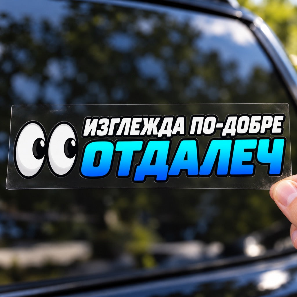 Ръка държи стикер „Изглежда по-добре отдалеч“ с две очи върху плоско прозрачно транспортно фолио