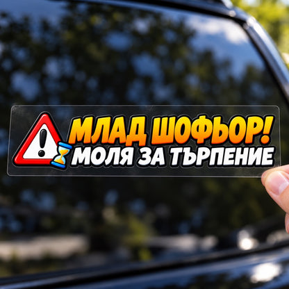 Ръка държи стикер „Млад шофьор! Моля за търпение“ върху плоско прозрачно транспортно фолио пред автомобил